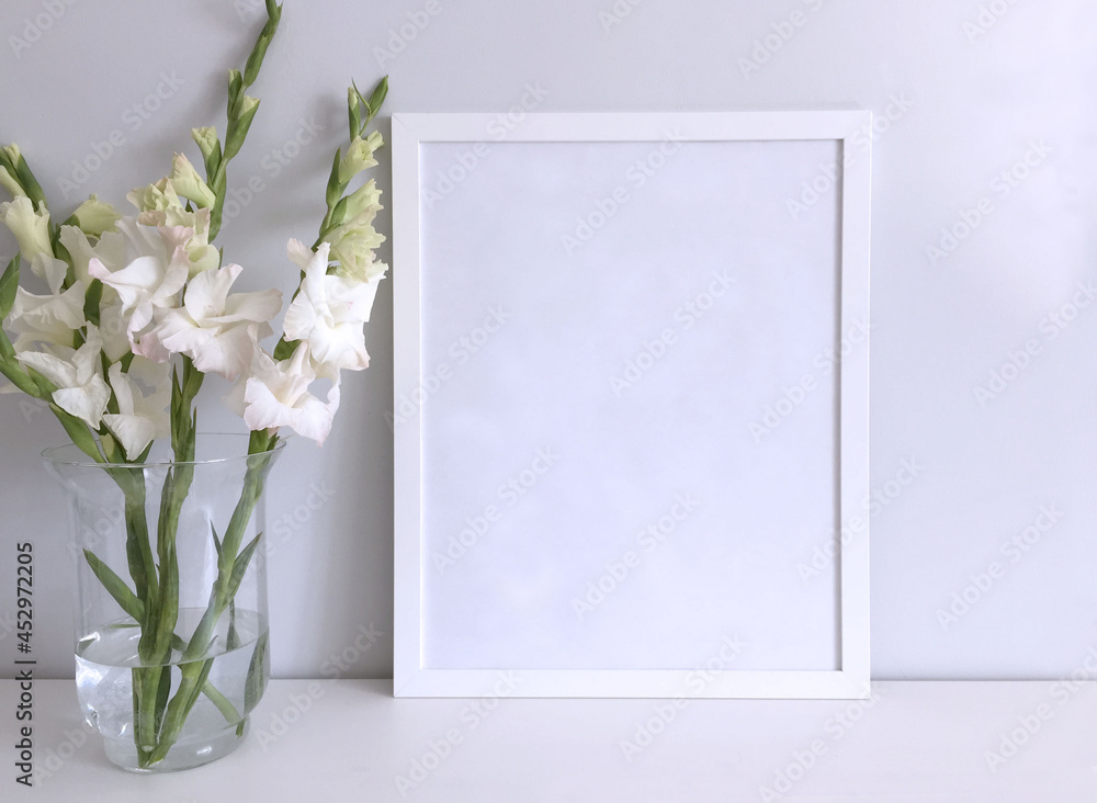 A frame is a template for a photo or picture on a white table surface with a bouquet of white gladioli in a large glass vase next to it. A frame is a template for a photo or picture on a white table surface with a bouquet of white gladioli in a large glass vase next to it.