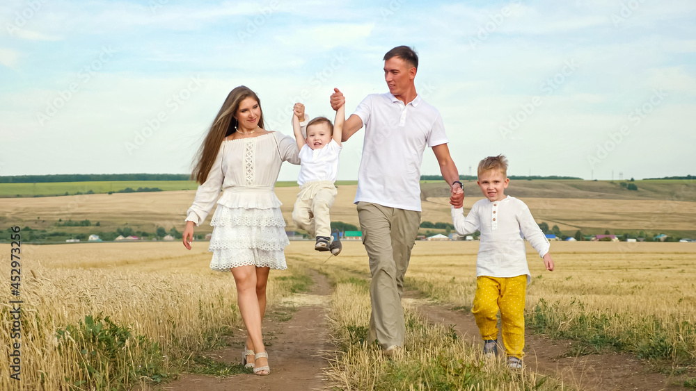 Fototapeta premium Family couple in white clothes walks joining hands with children and lifts toddler son in yellow wheat field in hot summer, against blue sky on horizon