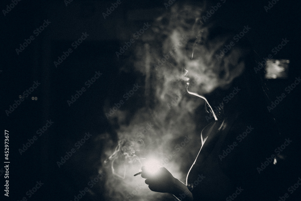 Young woman smokes cigarette in the dark with light spot ray. Black and white image with film grain