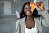 young african american woman in business suit smiling, covering eye with a slice of pizza.