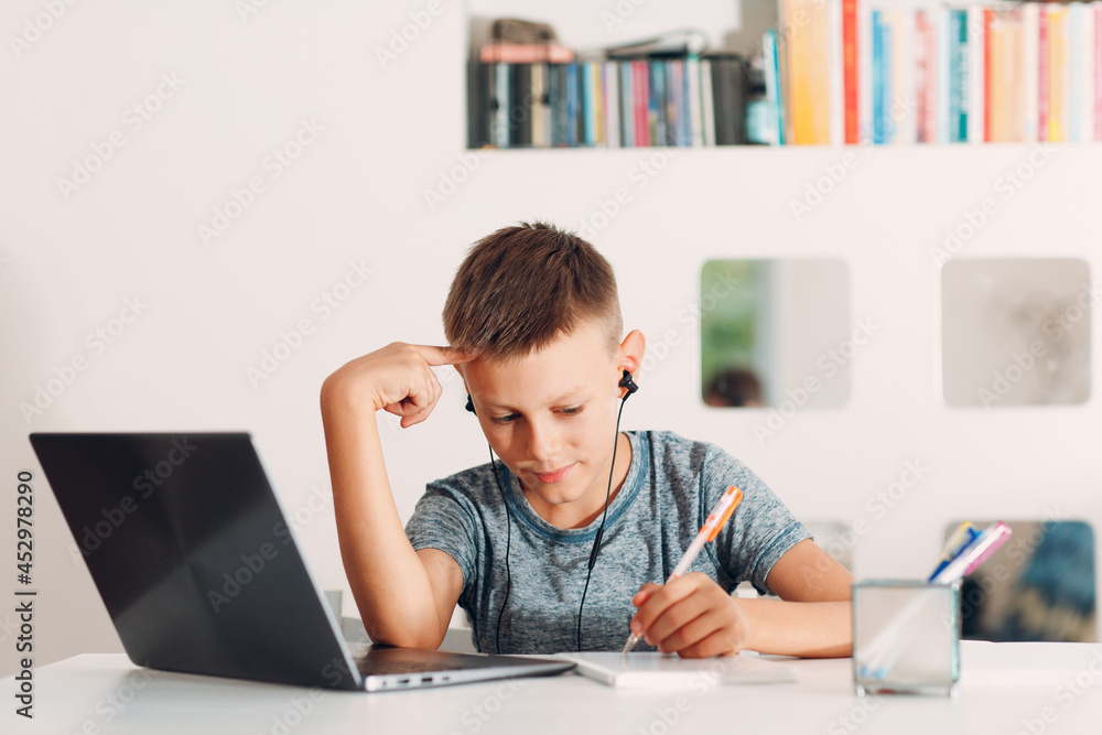 Young boy in headset sitting at table with laptop and preparing to school. Online education concept Young boy in headset sitting at table with laptop and preparing to school. Online education concept