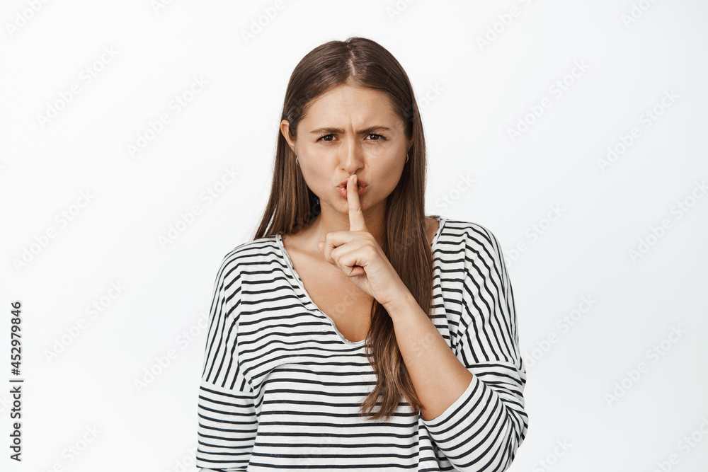 Angry young girl boss shushing, making shh hush gesture, press finger to lips in taboo gesture, telling to be quiet, white background Angry young girl boss shushing, making shh hush gesture, press finger to lips in taboo gesture, telling to be quiet, white background