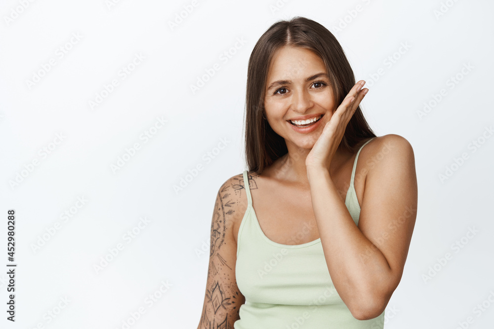 Beauty and skin care concept. Young woman laughs and smiles, touches her face, stands in tank top against white background Beauty and skin care concept. Young woman laughs and smiles, touches her face, stands in tank top against white background