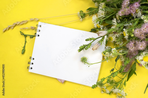 A notebook with a white blank sheet on a yellow background near a bouquet of wild flowers. Greeting card for greetings. Background photo