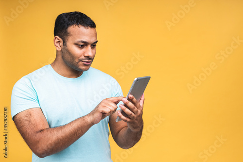 Using tablet. Happy winner! Young handsome african american indian man smiling holding tablet and playing games or using a booking app isolated over yellow background. © denis_vermenko