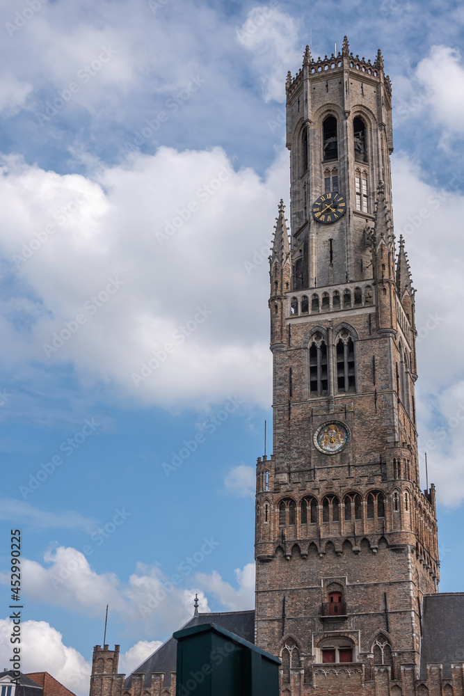 Brugge, Flanders, Belgium - August 3, 2021: Brown stone Tower of Halletoren Belfry against blue cloudscape. Brugge, Flanders, Belgium - August 3, 2021: Brown stone Tower of Halletoren Belfry against blue cloudscape.