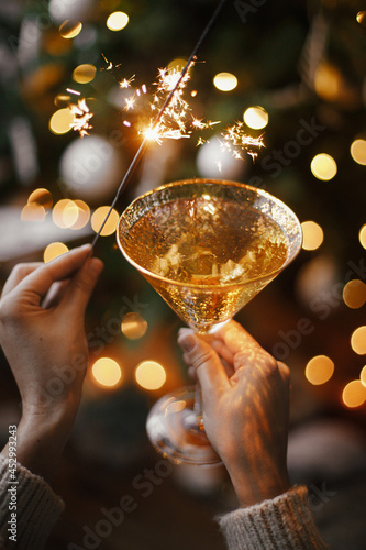 Happy New Year! Woman celebrating with firework light and champagne glass on background of christmas tree and glowing star. Hands holding burning sparkler and drink. Atmospheric moment