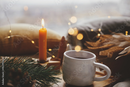 Warm cup of tea with christmas lights, stars, pine trees, candle and pillows on soft bed in scandinavian evening room. Cozy home. Atmospheric magic moment, winter hygge. Comfort and relax