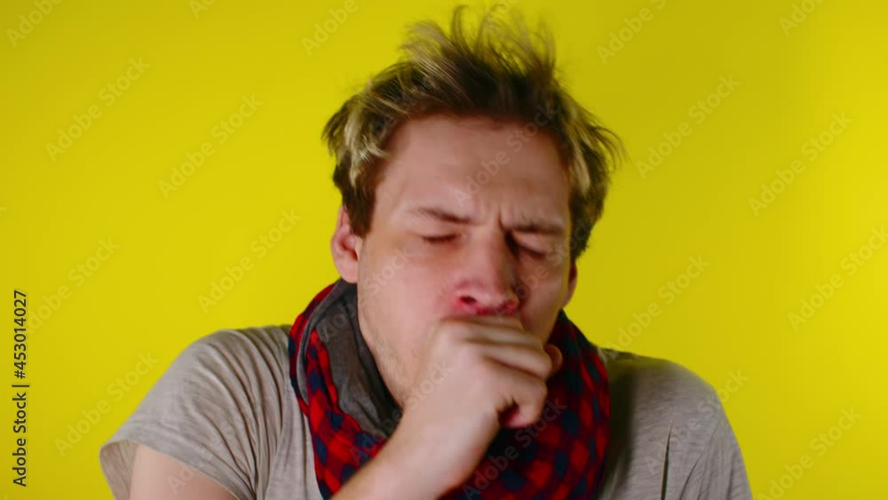 Young man wearing t-shirt and in a scarf, standing over isolated yellow background feeling unwell and coughing as symptom for cold or bronchitis. A man with a sore throat and flu symptoms. Young man wearing t-shirt and in a scarf, standing over isolated yellow background feeling unwell and coughing as symptom for cold or bronchitis. A man with a sore throat and flu symptoms.