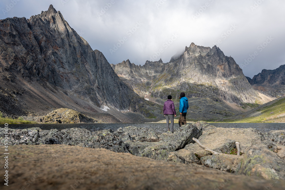 Two people, hikers, travellers standing beside Grizzly Lake in northern Canada during summertime wearing blue, purple clothing. 