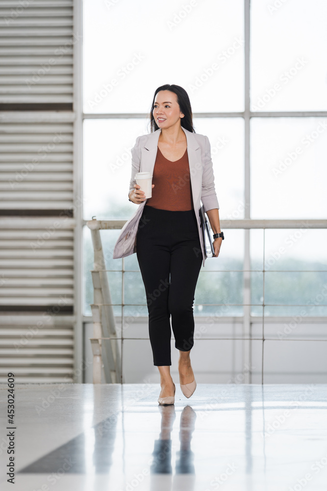 A happy successful Asian businesswoman holds a takeaway coffee cup, document file and tablet walks in the business building