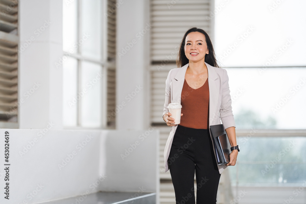 A happy successful Asian businesswoman holds a takeaway coffee cup, document file and tablet walks in the business building A happy successful Asian businesswoman holds a takeaway coffee cup, document file and tablet walks in the business building