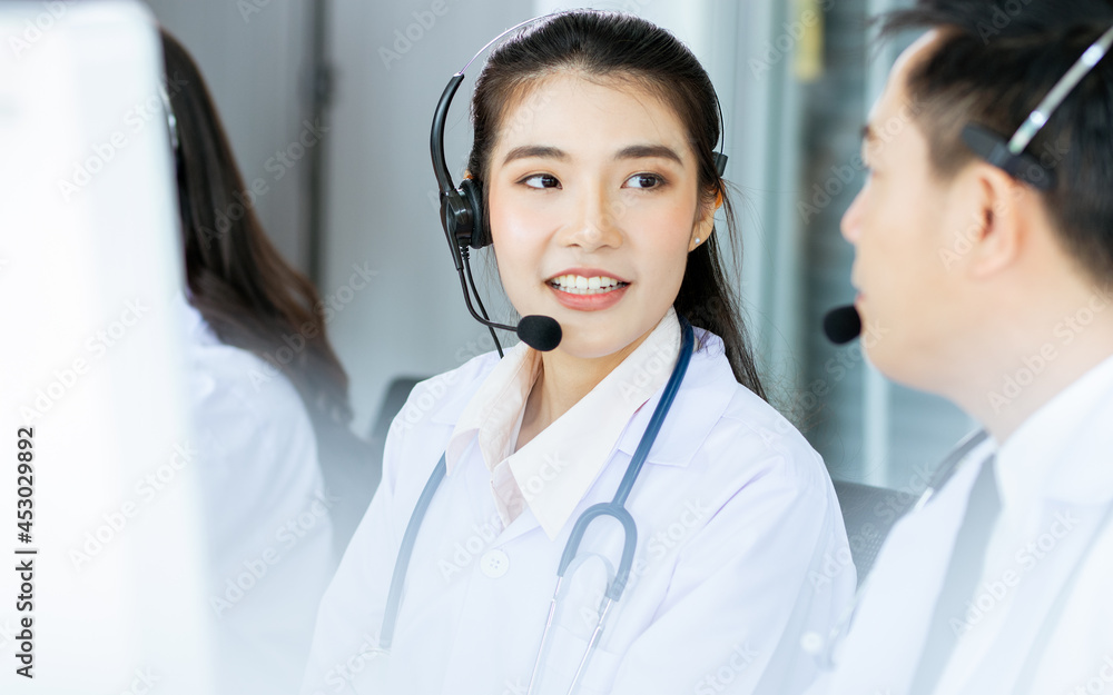 Selective candid Focus Asian beautiful female doctor smiling with headsets, sitting, monitoring and working with colleagues in hospital to advise online telemedicine to patients with service mind. Selective candid Focus Asian beautiful female doctor smiling with headsets, sitting, monitoring and working with colleagues in hospital to advise online telemedicine to patients with service mind.