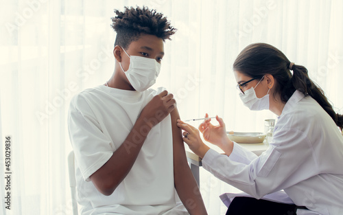 African teen man wearing face mask, getting vaccination to protect or prevent virus, getting scary vaccine injection while female caucasian doctor preparing syringe to vaccinate patient at hospital.