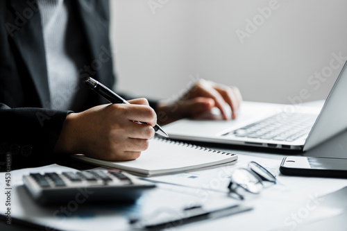 Close up of businesswomen or accountant using writing a notebook while working analytic business report on a laptop computer at the workplace, Planning financial and accounting concept.