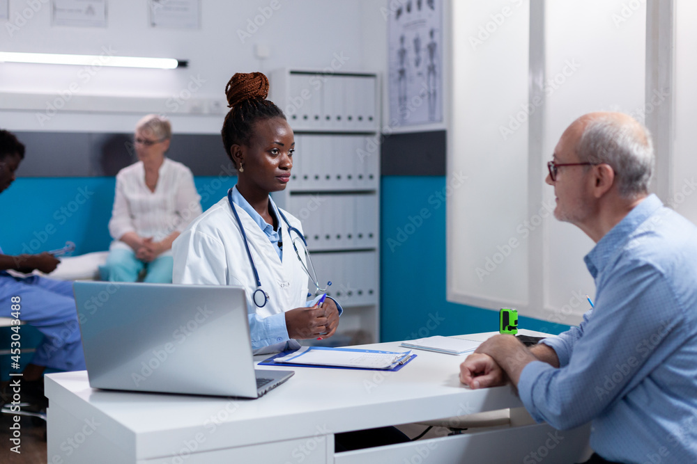 Black adult with medic occupation talking to senior patient sitting at desk in cabinet. African american doctor and sick old man having conversation about healthcare treatment and medicine Black adult with medic occupation talking to senior patient sitting at desk in cabinet. African american doctor and sick old man having conversation about healthcare treatment and medicine
