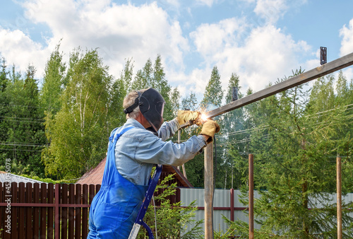 Man welder in a welding mask, construction uniform and protective gloves cooks metal on a street construction site. Construction of a pavilion, pergola near a country house on a summer day.
