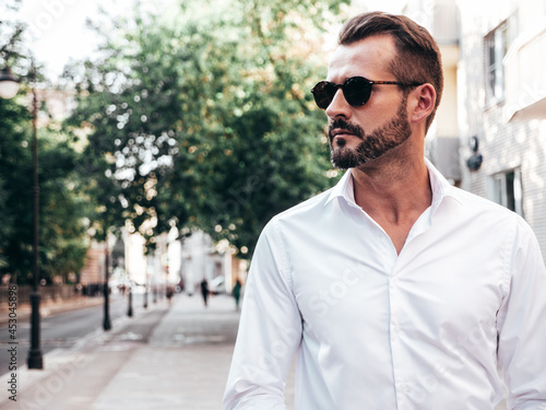 Portrait of handsome confident stylish hipster lambersexual model.Modern man dressed in white shirt and trousers. Fashion male posing in the street background in Europe city at sunset. In sunglasses