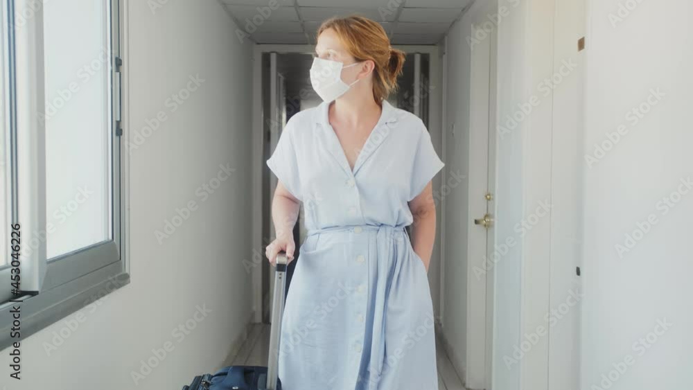 Vaccinated female tourist wearing protective medical mask in hotel after check in walks down corridor with suitcase. Vaccinated female tourist wearing protective medical mask in hotel after check in walks down corridor with suitcase.
