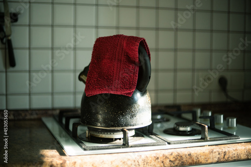 Interior of a light home kitchen on a summer morning. A black kettle stands on a gas stove with a red towel, a ray of sun is falling