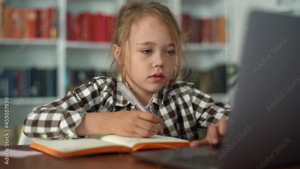 Close-up portrait of focused primary child school girl doing homework writing notes in paper notebook and looking at laptop. Adorable serious pupil schoolgirl e-learning online using computer app. Close-up portrait of focused primary child school girl doing homework writing notes in paper notebook and looking at laptop. Adorable serious pupil schoolgirl e-learning online using computer app.