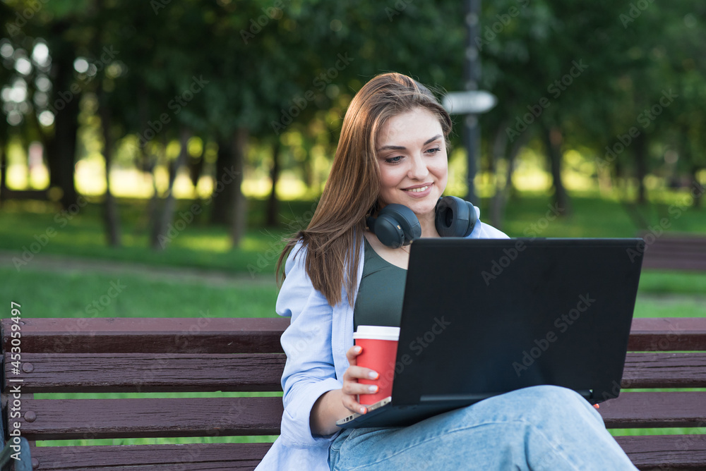 A young and attractive Caucasian girl in headphones sitting on a bench in the park. Using a laptop. Freelance, Network concept A young and attractive Caucasian girl in headphones sitting on a bench in the park. Using a laptop. Freelance, Network concept