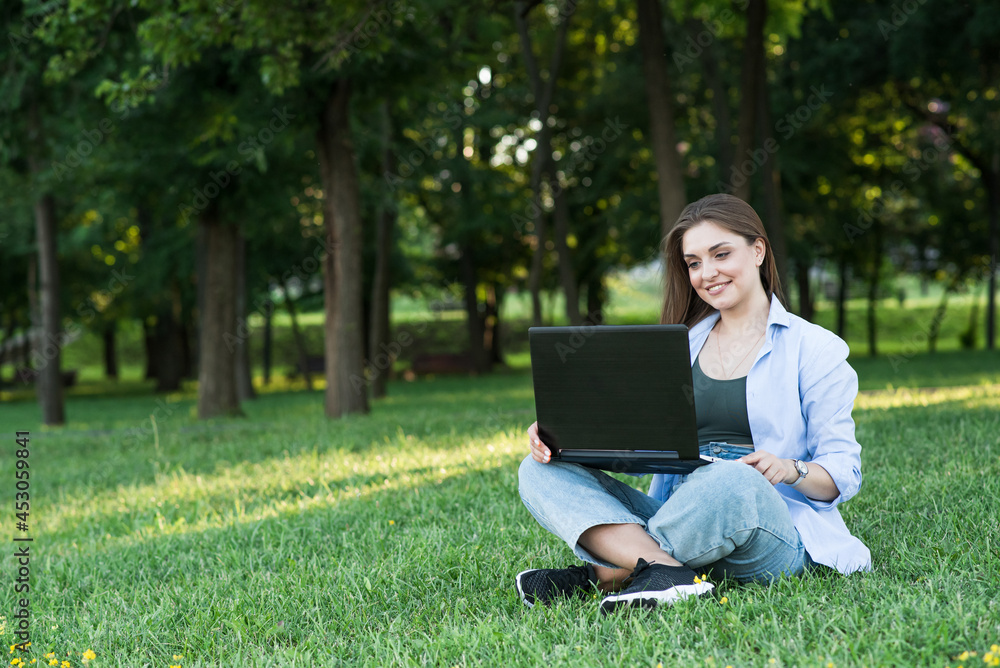 A young and attractive Caucasian girl sitting on the grass on the lawn and working. Using a laptop. Freelance and education concept. A young and attractive Caucasian girl sitting on the grass on the lawn and working. Using a laptop. Freelance and education concept.