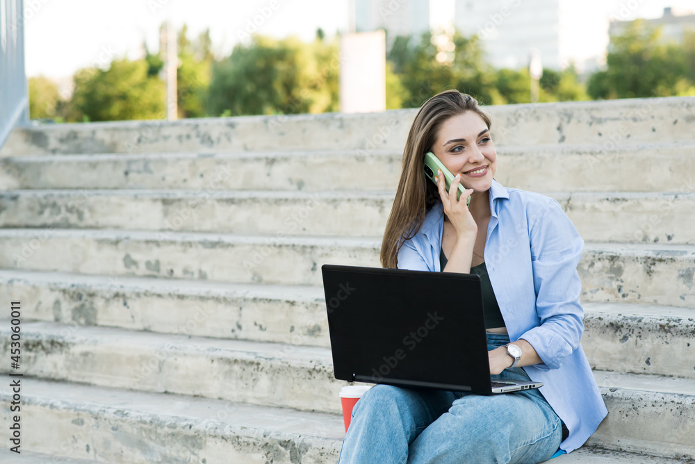 Happy and attractive caucasian girl is sitting with a laptop on the steps and talking on the phone. Distance working. Freelance concept Happy and attractive caucasian girl is sitting with a laptop on the steps and talking on the phone. Distance working. Freelance concept