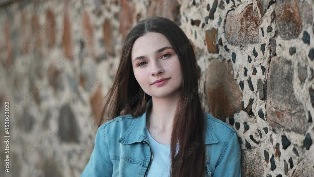 Portrait of a long-haired girl in a denim jacket against a stone wall.