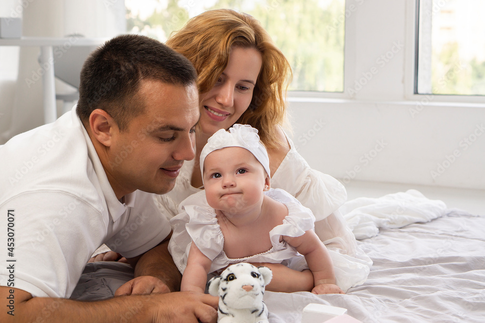 Happy family with girl baby having fun playing together on the floor at home. Background with copy space.Loving parents look at a child who smiles slyly.Little dreamer. Happy family with girl baby having fun playing together on the floor at home. Background with copy space.Loving parents look at a child who smiles slyly.Little dreamer.