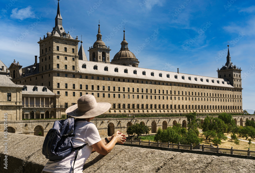 Mature woman with hat and small backpack contemplating the monument of the monastery of San Lorenzo de El Escorial, in summer and with blue sky and light white clouds.