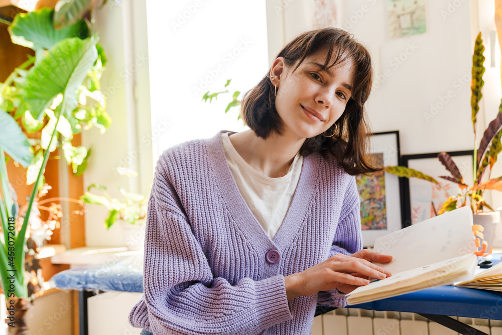 Young white woman looking into sketchbook while sitting indoors Young white woman looking into sketchbook while sitting indoors