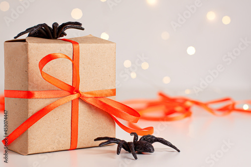 Close up of brown gift box with a orange satin ribbon bow and black horror spiders on bright blurred background with bokeh with copy space. Holiday autumn concept