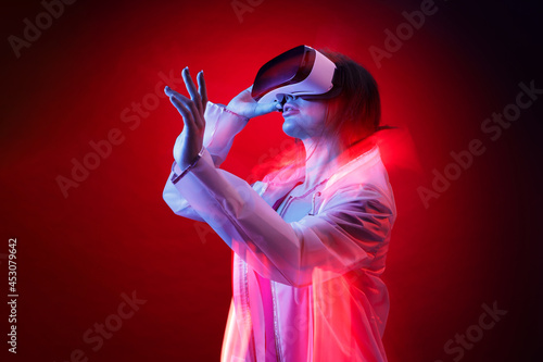 Woman in futuristic costume. Girl in glasses of virtual reality while touching air. Augmented reality game, future technology, AI concept. VR. Neon red light. © KDdesignphoto