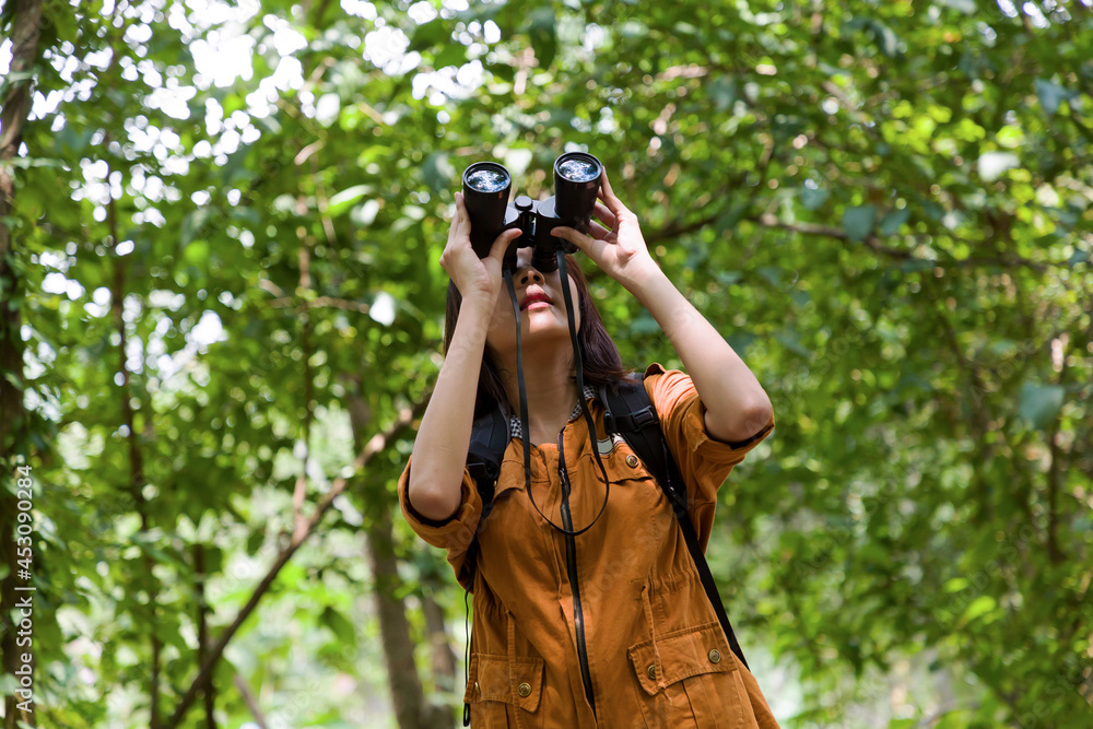 Portrait of Asian young woman hiker hiking with backpack standing holding binoculars looking for birds along forest trail