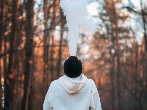 A young man in a white hoodie in an autumn park exhales a cloud of smoke