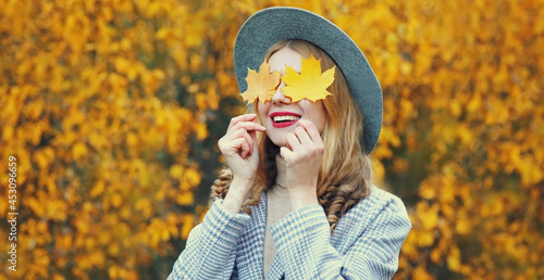 Autumn portrait of beautiful happy smiling woman with yellow maple leaves wearing a round hat in a park