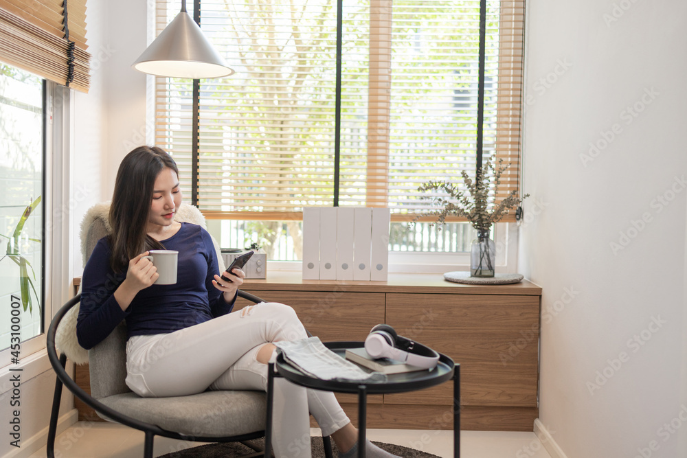 Work from home concept a confident woman sitting on a modern chair hold a cup of coffee and another hand typing a keyboard on her smart phone Work from home concept a confident woman sitting on a modern chair hold a cup of coffee and another hand typing a keyboard on her smart phone