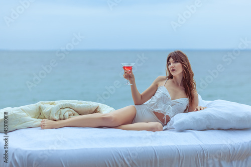Woman lies on a bed outdoors by the sea. She holds a glass of wine in her hand.
