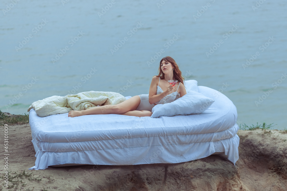 Bed with a beautiful woman is on a steep bank. Beautiful woman lies on a bed by the ocean.