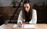 home office brunette girl writes something with a pen in a notebook lying on a light table. Adult female student studying at home remotely doing homework. Online education concept.