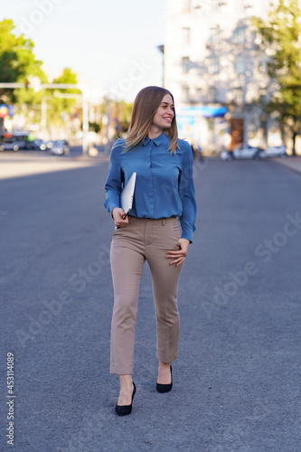 Blonde young woman smiling portrait wearing blue gentle shirt holding a laptop while walking