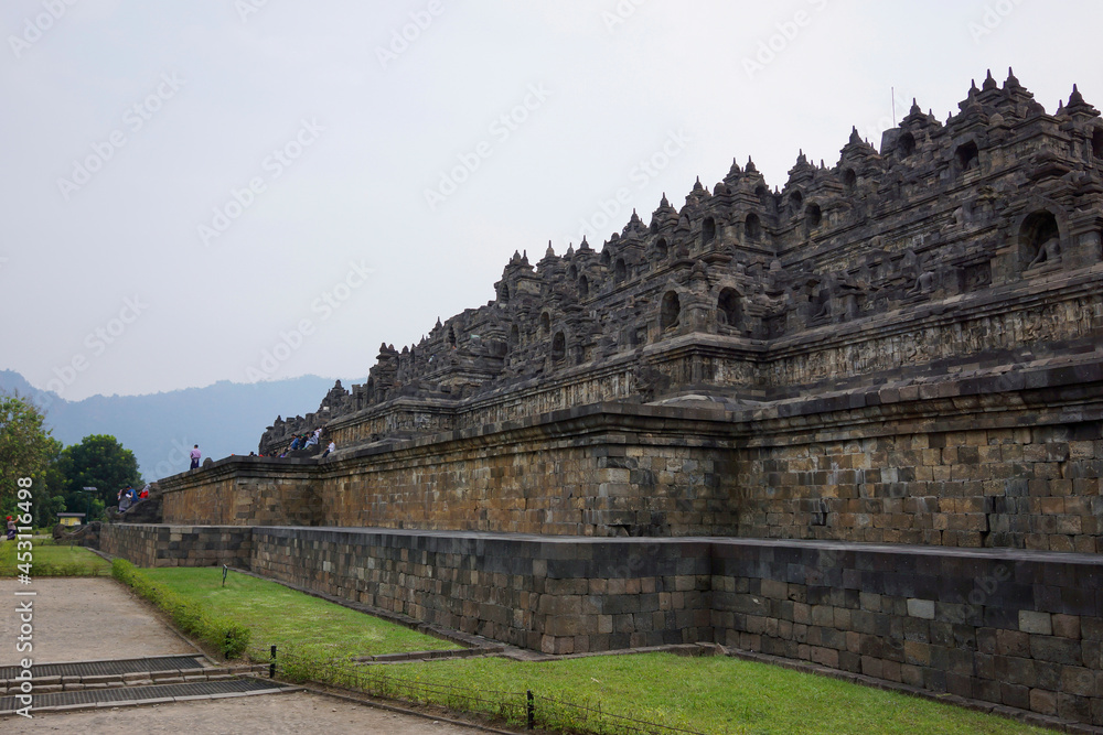 Exterior wall of ancient Borobudur temple view from the base of temple with mountain in background and clear blue sky. No people. Popular tourist and Buddhist pilgrimage destination.