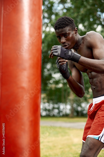 An african american boxer works with a punching bag, practicing punches with gloves, a black atleb boxer, trains in the city among the boxing grounds