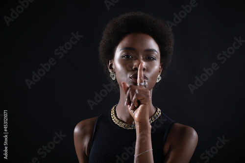 Closeup of beautiful young african american dark-skinned woman with finger on her lips showing silence gesture on black background. photo