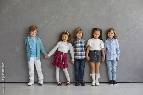 Portrait of cute and happy little boys and girls in casual clothes standing against the gray wall. Beautiful smiling and funny kids standing in a row holding hands. Childhood concept.