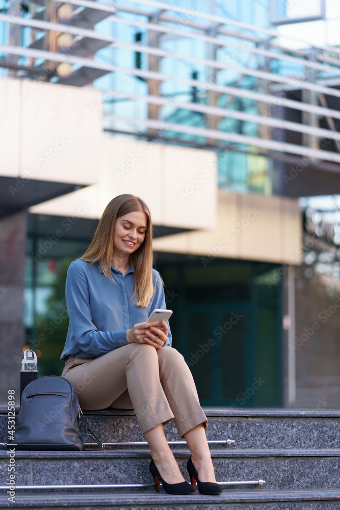 Young professional woman sitting on stair in front of glass building, talking on mobile phone