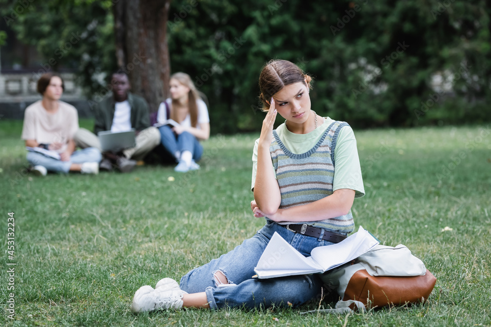 Fototapeta premium Upset student with notebook and backpack sitting on grass in park