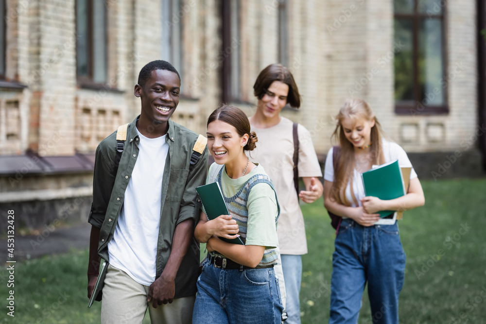 Cheerful interracial students with laptop and notebook walking near friends Cheerful interracial students with laptop and notebook walking near friends