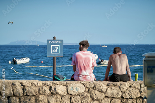 Fototapeta Naklejka Na Ścianę i Meble -  Sign with the inscription dog beach on a campsite in Krk in Croatia. Next to it two vacationers resting on a wall.